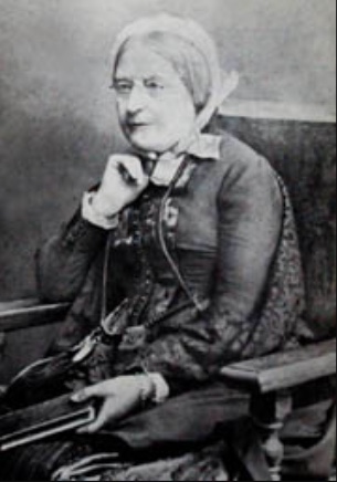 Maria Grey as a older woman sits in a chair holding a book. She wears glasses and a bonnet.