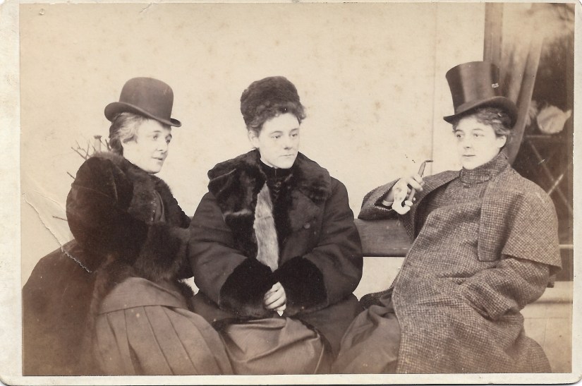 Cabinet card photograph from the 1890s of three upper class women seated on a bench. The woman on the left sits side on and is wearing a fur coat and riding or bowler hat. The woman in the middle also wears a fur coat and a small hat. The woman on the right is slightly reclined wearing a tweed Inverness Cape. She wears a top hat and is holding a smoking pipe.